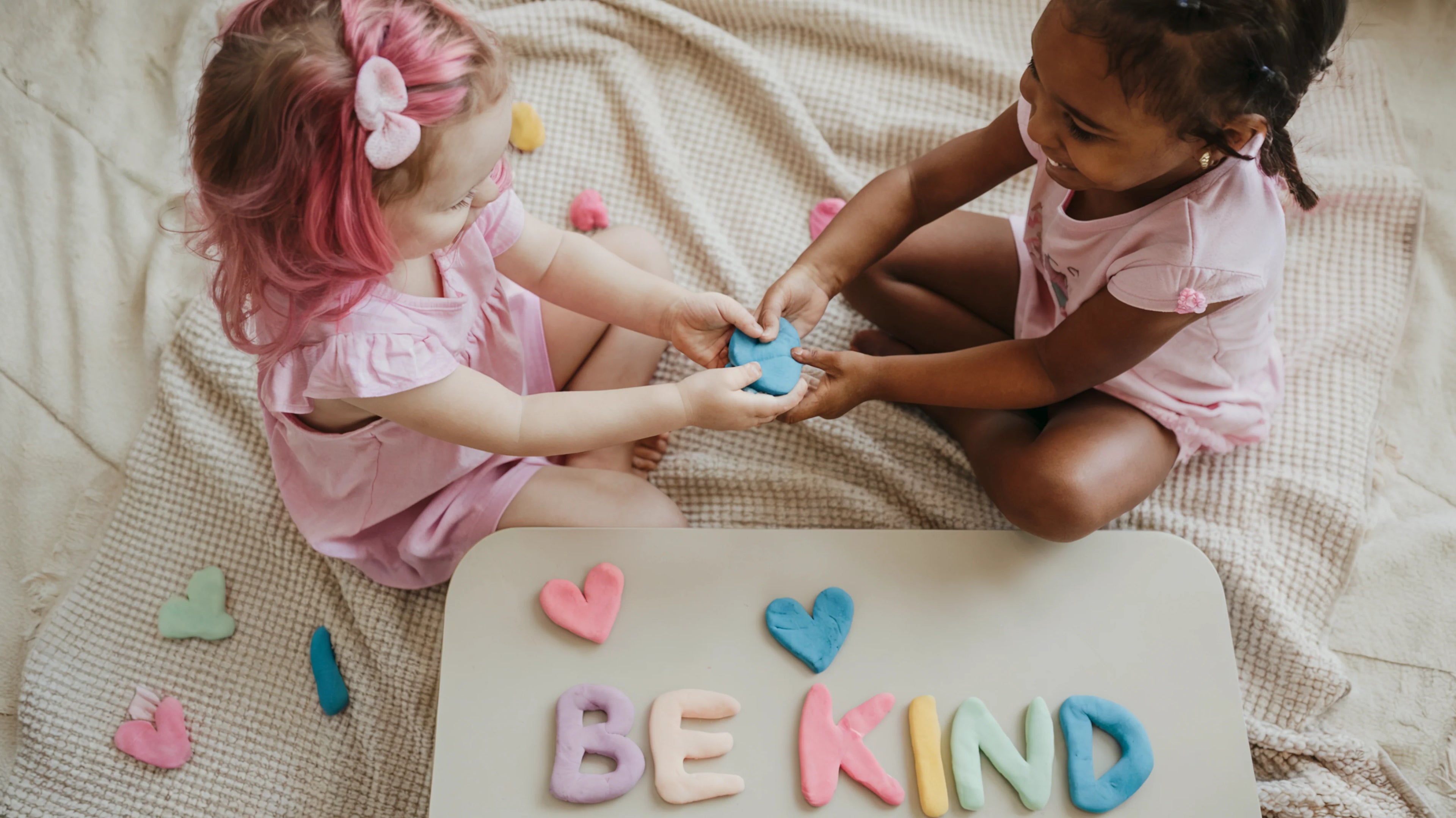 Two young girls playing with colorful heart-shaped blocks on a blanket, with 'BE KIND' spelled out in front of them.