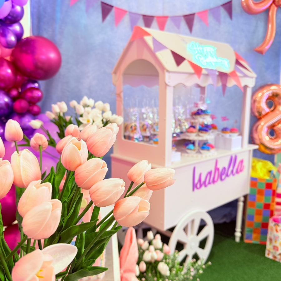 Decorative setup with flowers, balloons, and a candy cart named 'Isabella' for a celebration.