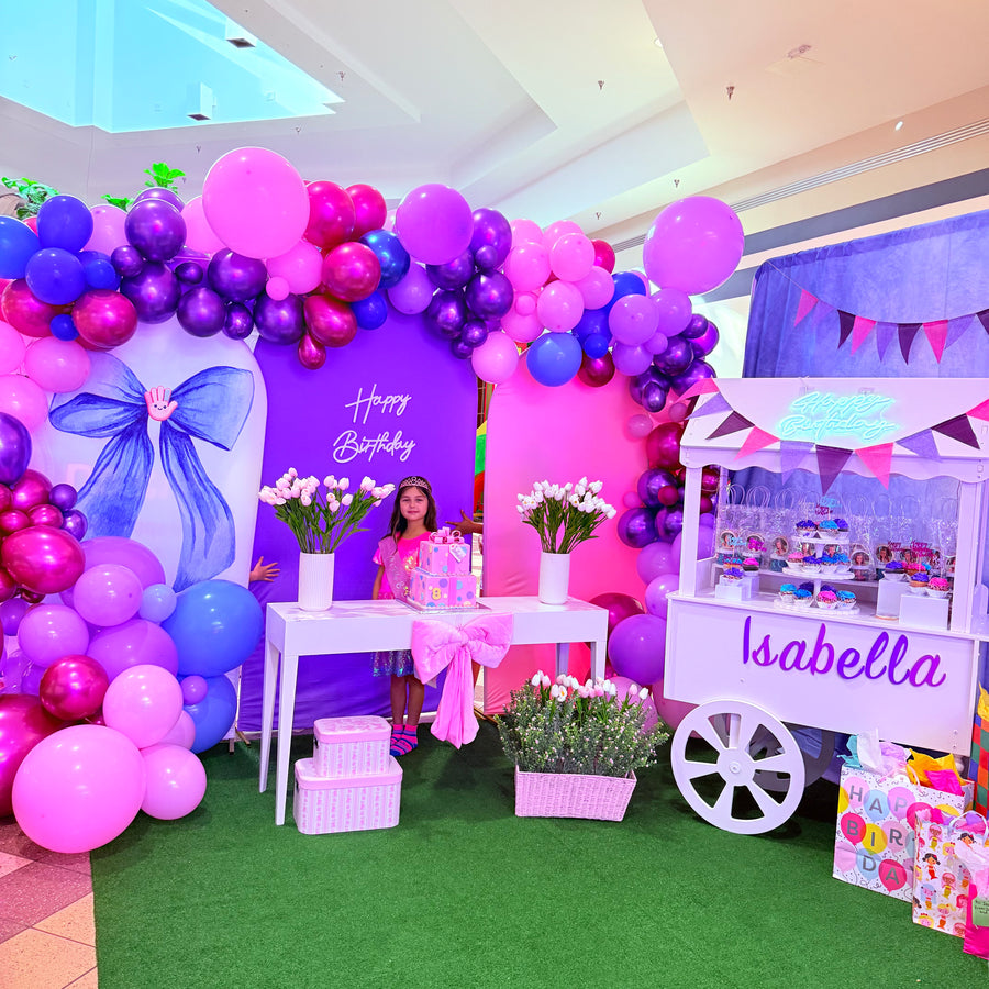 Decorated birthday party setup with balloons, flowers, and a cart named 'Isabella' in a mall.